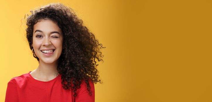 Lifestyle. Portrait of funny and cool sister with curly hair winking playfully having fun and foolind around showing tongue as playing with siblings adoring spend time with chidren over yellow background.