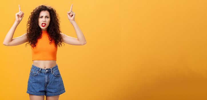 Indoor shot of displeased female client with curly stylish hairstyle. red lipstick in cool cropped top pointing up demanding explanation with confused and dissatisfied expression over orange background.
