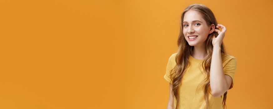 Cute and tender flirty young woman with cute wavy hairstyle flicking hair strand behind ear gazing at camera with timid sensual smile glancing with admiration at guy over orange background. Copy space