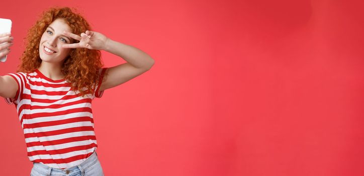 Hey followers how your summer holidays. Cheerful self-assured stylish fashionable redhead curly girl strike pose show peace victory gesture look smartphone screen taking selfie red background.