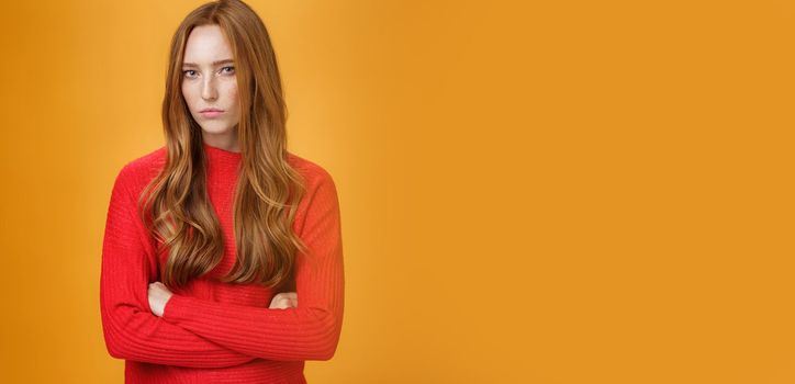 Suspicious intense and defensive ginger girl standing in passive-aggressive pose pouting and frowning looking with disbelief and disdain at camera, offended against orange background. Body language concept