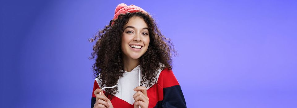 Close-up shot of carefree attractive feminine girl with curly hair in beanie playing with sweatshirt as posing over blue background, smiling at camera having fun and positive attitude.