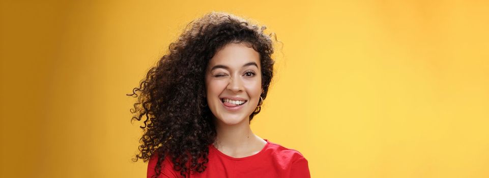 Lifestyle. Portrait of funny and cool sister with curly hair winking playfully having fun and foolind around showing tongue as playing with siblings adoring spend time with chidren over yellow background.
