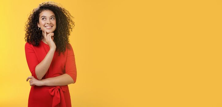 Girl having interesting idea, smiling as feeling confident plan might work. Charming happy woman with curly hair in red dress gazing at upper left corner thoughtful, thinking over yellow wall.