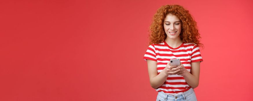 Time check social media feed. Good-looking relaxed happy fashionable redhead woman curly hairstyle hold smartphone look phone screen amused smiling delighted texting friend throw party asap.
