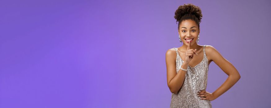 Excited joyful african-american young 20s woman in silver dress telling secret gossiping showing shh gesture say shush smiling gladly wanna make surprise, hold hand waist index finger on mouth.