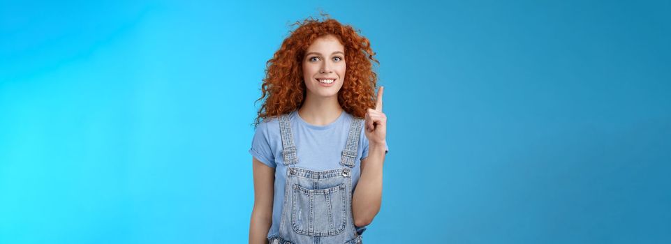 Lifestyle. Cheerful cute energized redhead curly girl wear denim overalls book summer vacation trip offer good offer link pointing raised index finger up show uppwards advertisement smiling silly.