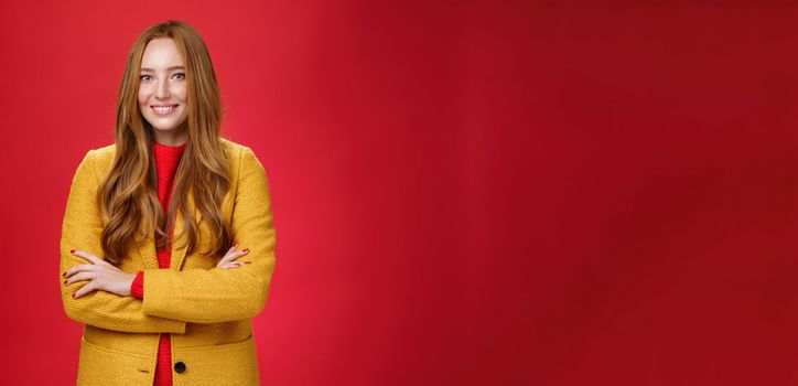 Lifestyle. Charming successful young female in yellow autumn coat smiling broadly holding hands crossed on body in confident self-assured pose standing delighted and happy over red background.