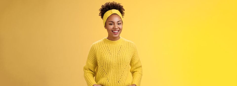 Friendly outgoing relaxed young african-american beautiful girl headband forehead in sweater hold hands pockets smiling broadly have fun talking interesting communication, standing yellow wall.