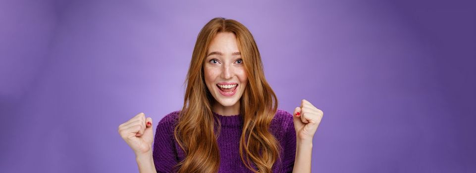 Optimistic, lucky young attractive female student winning trip around european smiling broadly from success and delight clench fists in triumph and celebration gesture, happy to win over purple wall.