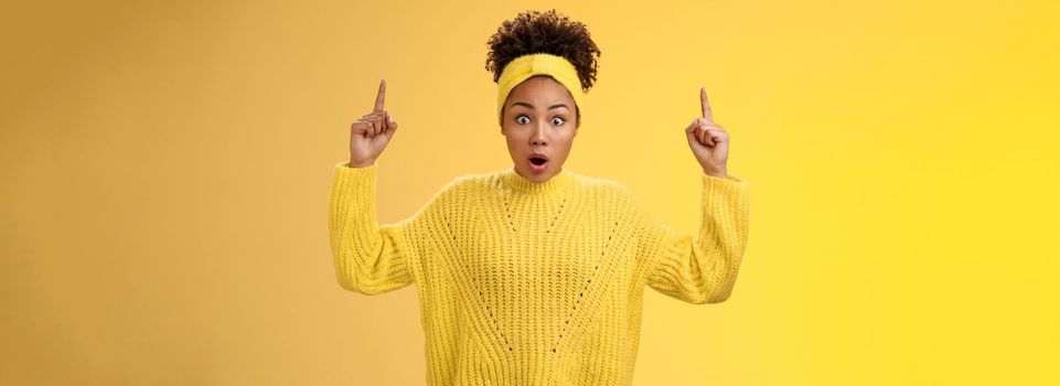 Shocked surprised emotive african-american female student drop jaw widen eyes learn astonishing news pointing raised fingers up gasping concerned impressed, speechless standing yellow background.