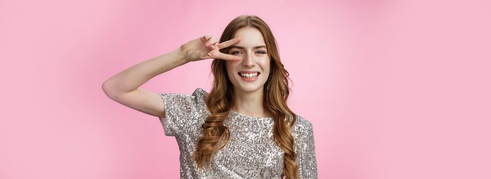 Positive carefree lucky charming glamour woman curly hairstyle party outfit having fun showing victory peace gesture dancing disco smiling broadly enjoying awesome atmosphere, pink background.