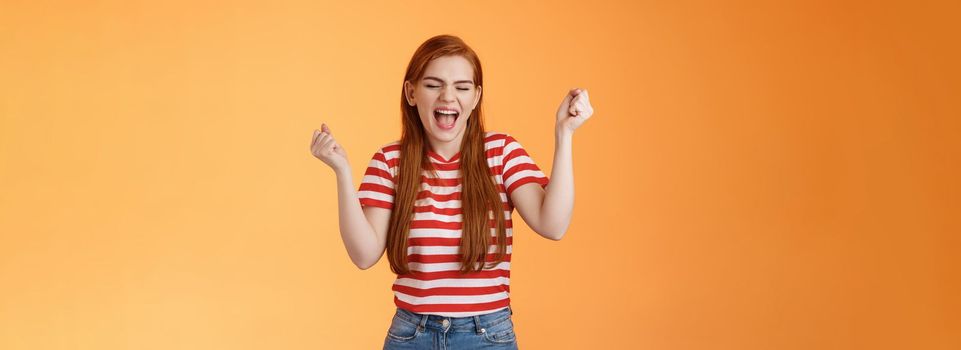 Luck cute redhead girl, make fist pump cheerfully yell yes, close eyes, winning lottery, feeling amazed, celebrate success and victory, stand orange background, triumphing, rejoicing good news.