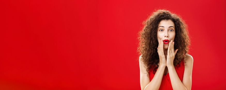 Charmed and intrigued curious good-looking rich woman in elegant dress with make-up and curly hairstyle folding lips in wow sign holding hands on face and smiling from excitement over red background. Emotions concept