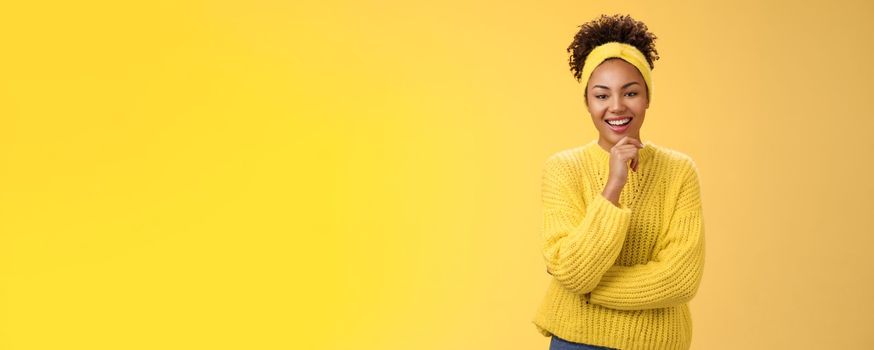 Interested excited enthusiastic good-looking female entrepreneur stylish sweater headband afro hairstyle touching chin thoughtful smiling liking project idea standing yellow background intrigued.