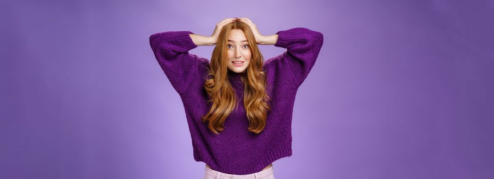 Cute european redhead 20s woman acting nervous, holding hands on head raising eyebrows in worry and anxiety, panicking being troubled think up plan posing over purple background. Copy space