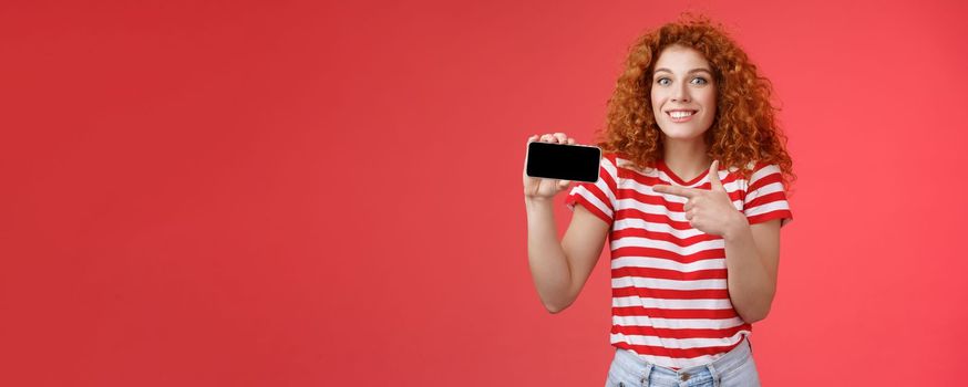 Excited happy good-looking european redhead curly girl amazed showing personal achievement score smartphone game show phone screen pointing display discuss awesome gameplay smiling delighted.