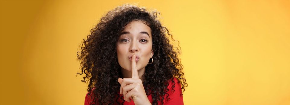 Shh please not tell secret. Cute and tender woman with curly hairstyle gently shushing with index finger on folded lips asking keep promise and remain silent, making surprise over yellow wall.