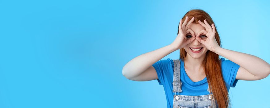 Playful cheerful redhead european woman, long nature red hairstyle, show okay finger glasses look through ok signs, smiling broadly funny grimace, stand blue background carefree enjoy summer.