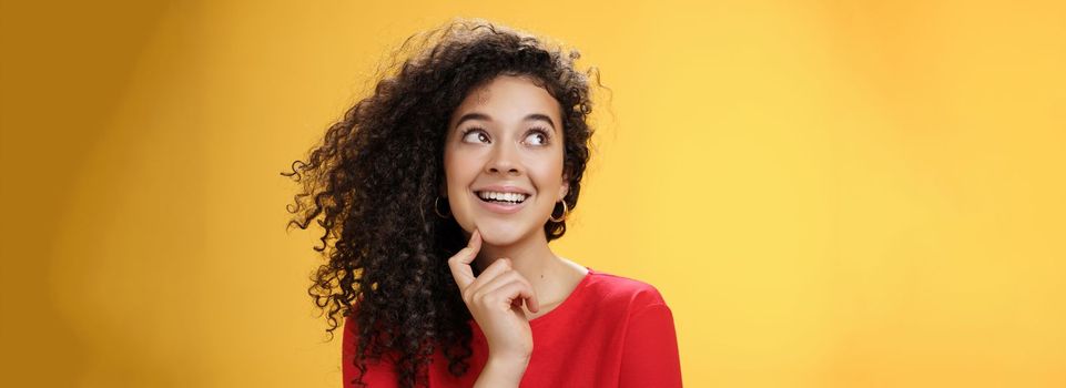 Dreamy girl making assumptions what gets as christmas present looking curious and happy at upper left corner, thinking, touching chin, standing thoughtful with desire and wish in mind over yellow wall.