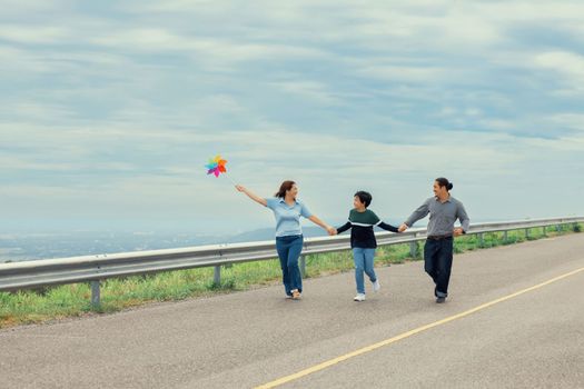 Progressive happy family vacation and carefree day concept. Young parents mother father and son run along and flying kite together road with enjoy natural scenic on scenery and clear sky background.