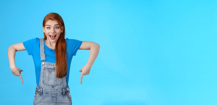 Amused wondered excited cute 20s european woman, pointing down, bottom copy space, look amazed admiration camera, describe amazing product, recommend site, stand blue background. Copy space