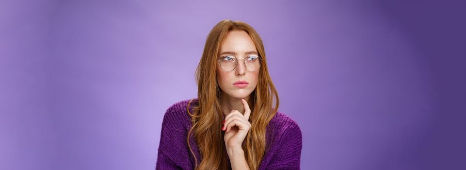 Girlfriend solving puzzle like detective making assumptions focusing on problem squinting looking left determined and thoughtful wearing glasses touching jaw while thinking over purple background.