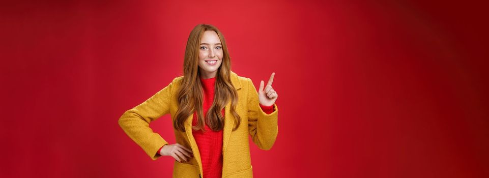 Charming young female in yellow coat holding hand on waist and pointing at upper left corner, smiling broadly as showing or promoting interesting proposal over red background, in good mood.