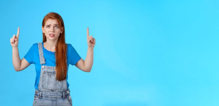 Uneasy worried cute redhead female coworker feel unsure insecure, grimacing displeased intense worried expression, look pointing up, top copy space, have hesitations, stand blue background.