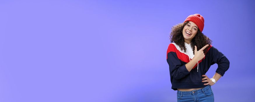 Studio shot of enthusiastic charming curly-haired woman inviting to play in snow with her pointing at upper right corner tilting head joyfully and smiling, holding hand on waist over blue wall. Emotions and weather concept