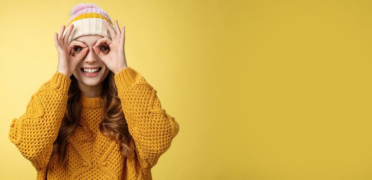 Surprised amused interested attractive young european woman wearing corduroy hat knitted sweater widen eyes curious show binocular glasses made fingers, smiling amazed, yellow background.