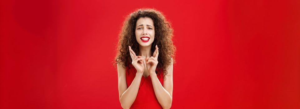 Concerned insecure hopeful woman. with curly hair in red lipstick and stylish dress clenching teeth frowning crossing fingers for good luck praying being in trouble and wishing deal went okay.