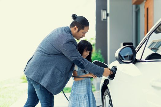 Progressive father and daughter plugs EV charger from home charging station to electric vehicle. Future eco-friendly car with EV cars powered by renewable source of clean energy.