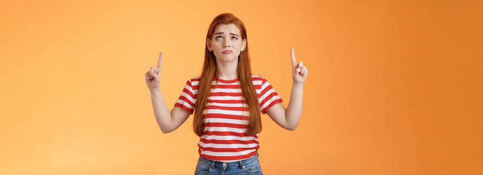 Hesitant upset cute redhead woman weighing choices look doubtful uncertain, frowning thoughtful, ponder decision, look pointing up unsure, have doubts, stand insecure orange background.