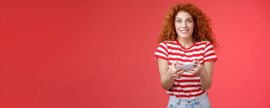Lifestyle. Excited happy cute redhead curly-haired girlfriend eager start learn how play boyfriend game look amused cheerful camera smiling joyful hold horizontal smartphone load app red background.