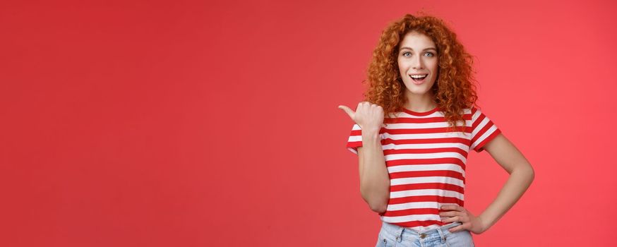 Lifestyle. Sassy good-looking emotive happy smiling redhead european female curly hairstyle pointing thumb left grin assertive cheeky hold hand waist directing promo advertising offer red background.