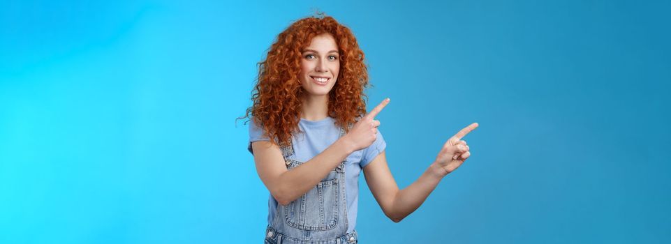 Cheerful upbeat positive summer girl redhead curly hairstyle wear denim overalls getting ready beach vacation pointing upper left corner promote store smiling delighted enjoy holidays.