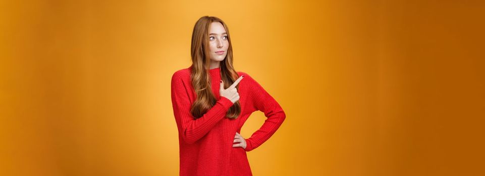 Portrait of glamour and stylish redhead female entrepreneur designing and leading new project pointing at upper left corner as gazing thoughtful and focused on job, managing business over orange wall.