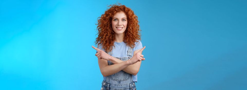 Which way. Charismatic attractive happy smiling redhead curly girl pointing curious sideways cross arms body indicating left right intrigued what choose making decision standing blue background.