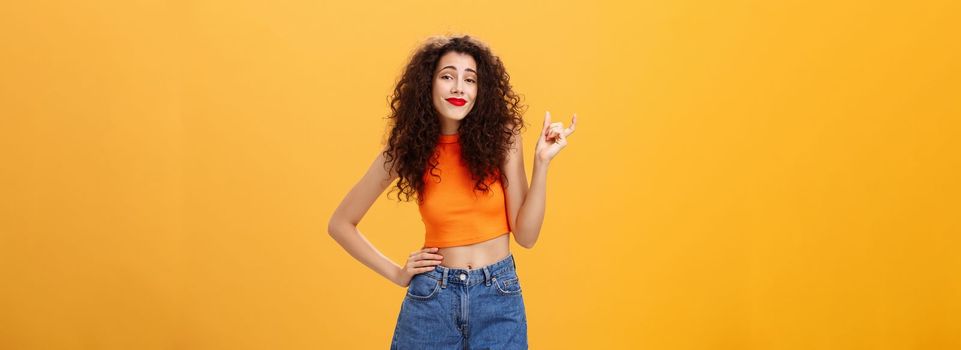 Girl needs bigger size to impress. Carefree picky stylish urban female with curly hairstyle red lipstick in cropped top shaping small or little thing with fingers smirking unimpressed over orange wall.