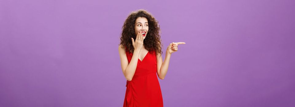 Girl chuckles over clumsy guy. making mistake during party covering smile with palm gazing and pointing left while gossiping with girlfriends joyfully standing in elegant red dress over purple wall.