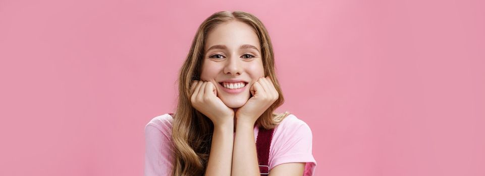 Lifestyle. Cute feminine and tender nice girl withtattoo and wavy hair leaning head on hands as if listening amusing story gazing with broad smile and admiration gaze being dreamy over pink background.