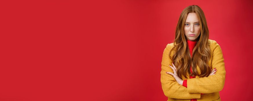 Offended and angry redhead girlfriend expressing dismay and hate as being waiting too long crossing hands over chest in defensive and aggressive pose, pouting looking with contempt over red background.