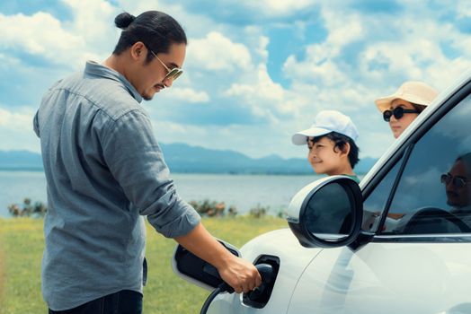 Concept of progressive happy family with electric vehicle enjoy their time at natural outdoor scenic with lake, greenfield and cloudscape background. Electric vehicle driven by green renewable energy.