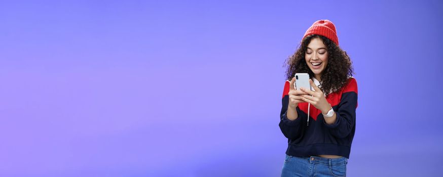 Lifestyle. Joyful and sociable stylish caucasian woman with curls in warm beanie having fun laughing and chuckling as using smartphone typing funny message looking at mobile phone screen happily over blue wall.