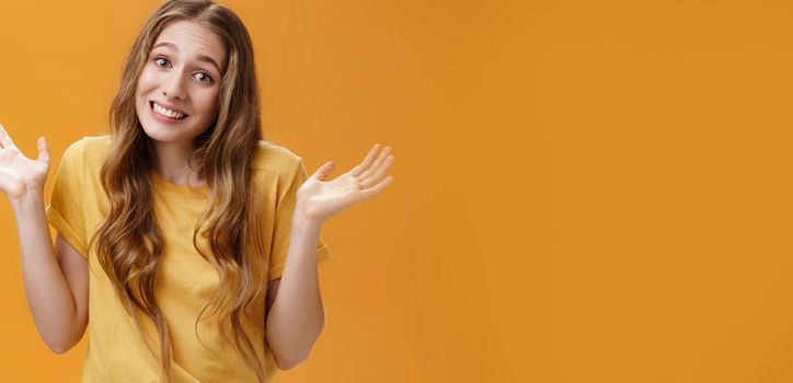 Girl stay out of business shrugging with hands raised and spread aside, silly sorry smile posing over orange background unaware and confused in casual t-shirt having no idea about topic.