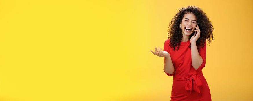 Portrait of joyful charming european female in red dress with curly hair laughing out loud as gossiping with friend via smartphone close eyes as giggling gesturing, hearing joke through mobile phone.