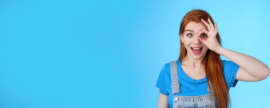 Dreamy curious attractive cheerful redhead woman surprised, look fascinated camera look through fingers okay sign, stand amused, gaze admiration blue background, check out promo. Copy space