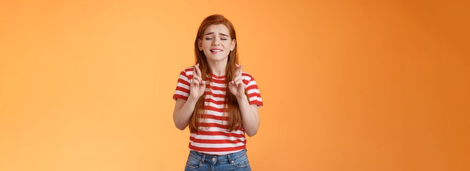 Hopeful cute redhead woman praying results be okay, faithfully cross fingers good luck, bite lip nervously believe she win, close eyes supplicating, plead lord dream come true, orange background.