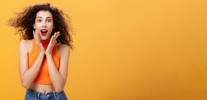 Waist-up shot of charmed enthusiastic attractive female with curly hairstyle in cropped top holding palms on cheeks and smiling astonished and surprised gazing with amazement over orange background. Copy space